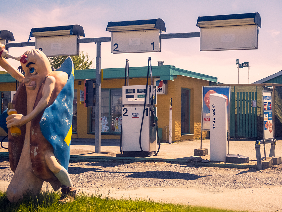 Udendørs tankstation med pumpeenheder, skiltet “Car Wash” og en stor oppustelig figur ved solnedgang.