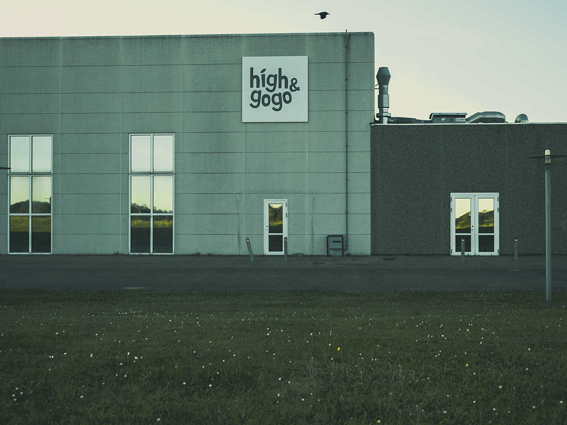 Grå industribygning med skiltet “high&gogo”, vinduer, dør og græsplæne foran i aftensol.