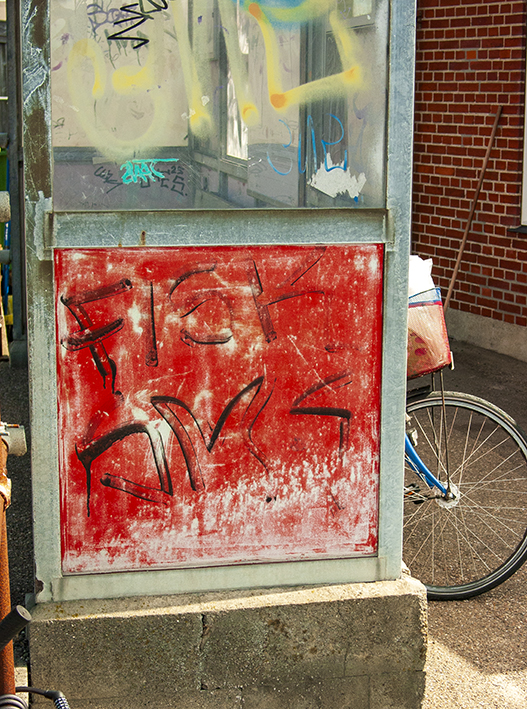 Rød graffiti-malet metalplade i en muråbning ved murstensvæg, cykel og skraldespand i baggrunden.